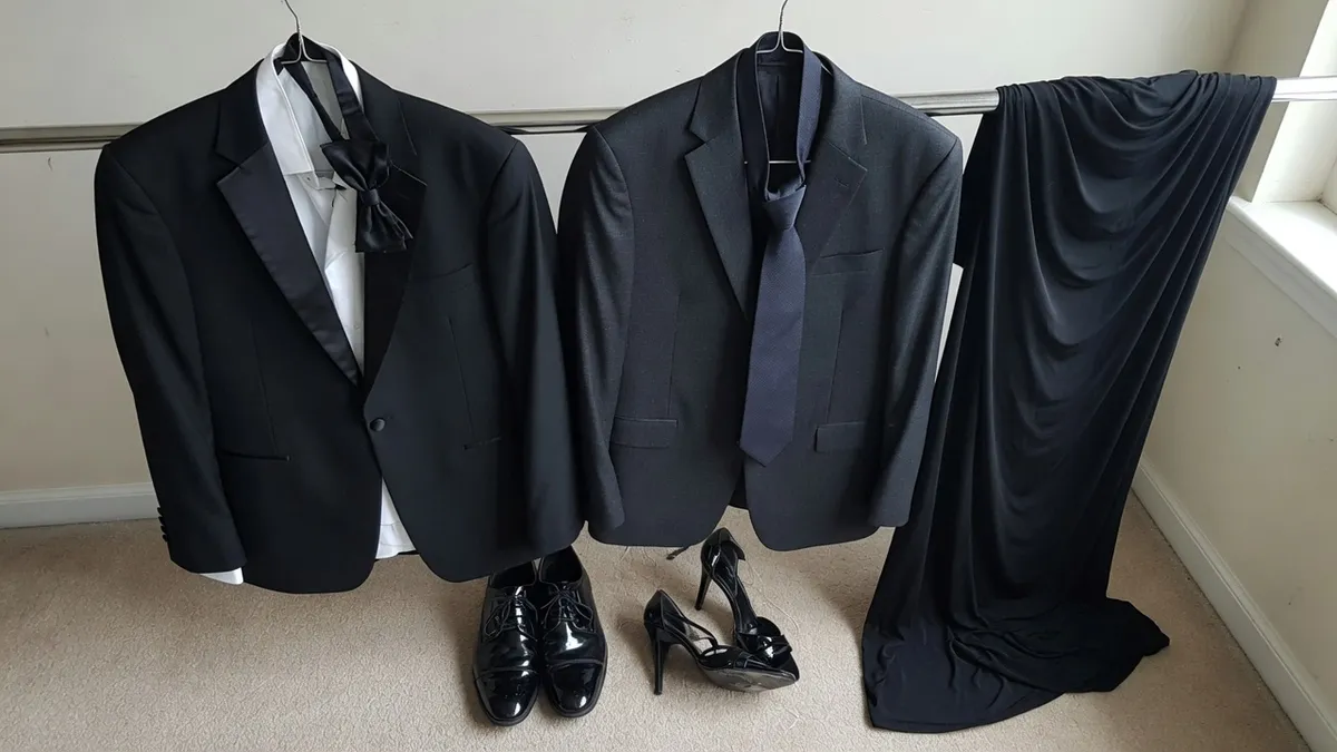 A black tuxedo with a bow tie and a charcoal suit with a dark tie hanging side by side on a rail, a long black gown beside them, patent shoes and heels below